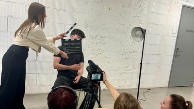 Mehrere Schüler:innen bei einem Filmdreh in einem hellen Raum. Eine Person hält eine Filmklappe vor einen sitzenden Schüler, während eine Kamera auf einem Stativ auf ihn gerichtet ist. Weitere Schüler:innen sitzen rund um die Kamera und begleiten die Aufnahme.