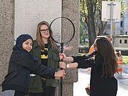 Gruppe von drei Mädchen auf der Straße. Sie alle halten mit ihren Händen eine Stange die am oberen Ende einen großen Ring hat.