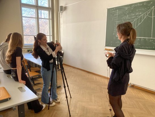 Schüler:innen in einem Klassenraum bei Dreharbeiten: Eine Schülerin bedient eine Kamera auf einem Stativ, eine weitere Person steht vor einer Tafel.