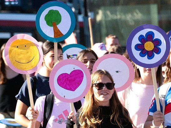 Schüler:innen bei einer Aktion im öffentlichen Raum mit selbst gestalteten, bunten Schildern mit Symbolen wie Smiley, Baum, Herz und Blume.