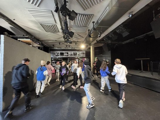 Schüler:innen bewegen sich bei einer Theaterprobe durch einen Bühnenraum mit schwarzem Boden und Bühnentechnik.