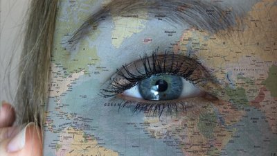 Nahaufnahme eines Auges, über das eine Weltkarte gelegt ist. Europa, Afrika sowie Teile Nord- und Südamerikas sind auf Gesicht und Augenpartie zu erkennen. Im Hintergrund sind Haare und ein Teil einer Hand sichtbar.
