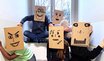 Mehrere Schüler:innen sitzen und stehen vor einem Fenster und halten selbst gestaltete Kartonmasken mit gezeichneten Gesichtern vor ihre Köpfe.