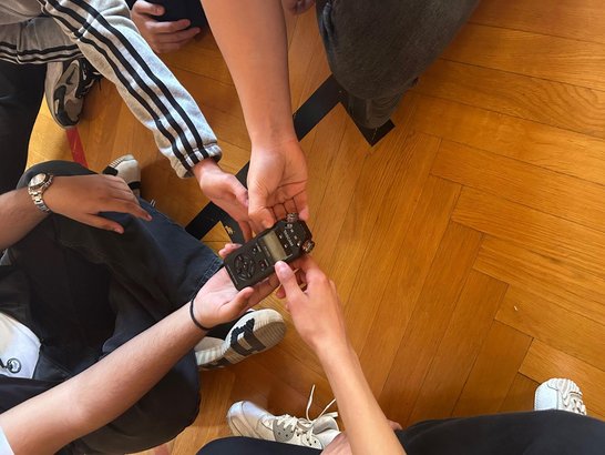 Mehrere Schüler:innen sitzen auf einem Holzboden und bedienen gemeinsam ein tragbares Audioaufnahmegerät.
