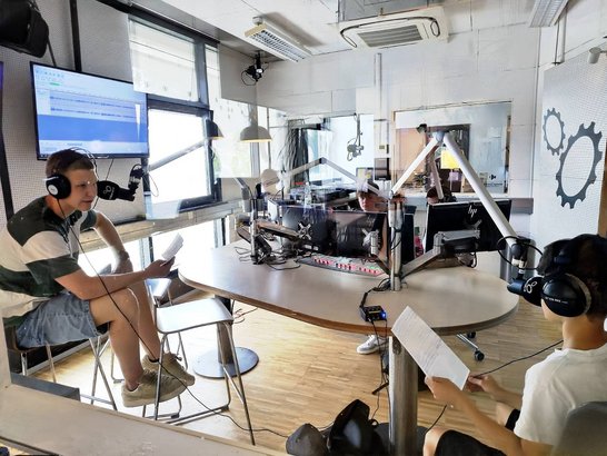 Mehrere Schüler:innen sitzen mit Kopfhörern und Manuskripten in einem Radiostudio vor Mikrofonen und produzieren gemeinsam eine Sendung.