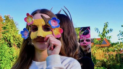 Zwei Schüler:innen stehen im Freien und halten selbst gestaltete, farbenfrohe Masken vor ihre Gesichter; eine weitere Maske ist in einem Spiegel zu sehen.
