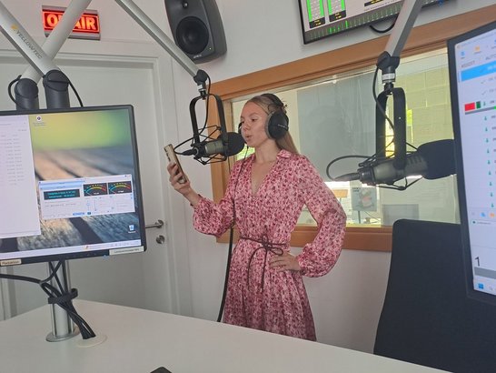 Eine Schülerin steht mit Kopfhörern vor einem Mikrofon in einem Radiostudio und liest von ihrem Smartphone ab.