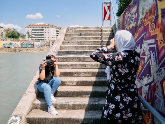Eine Person fotografiert eine weitere Person vor einer mit Graffiti gestalteten Mauer an einer Treppe entlang eines Gewässers.