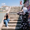 Eine Person fotografiert eine weitere Person vor einer mit Graffiti gestalteten Mauer an einer Treppe entlang eines Gewässers.