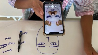 Eine Person hält ein Smartphone über eine Zeichnung. Die Kamera verbindet gezeichnete Gesichtszüge mit realen Objekten zu einer interaktiven digitalen Darstellung.