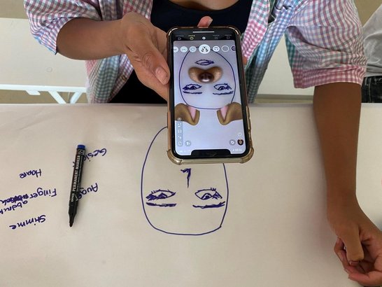 Eine Person hält ein Smartphone über eine Zeichnung. Die Kamera verbindet gezeichnete Gesichtszüge mit realen Objekten zu einer interaktiven digitalen Darstellung.