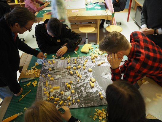 Kinder sitzen am Boden schneiden Häuser aus Schaumstoff um diese auf eine Stadtkarte zu platzieren.