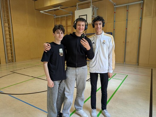 drei Schüler in Turnsaal, zwei mit Kopfhöhrer, einer mit Diktiergerät