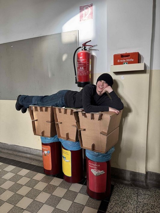 Foto von einem Schüler der auf 3 Hockern aus Karton liegt, die wiederum auf 2 Mistkübel stehen.