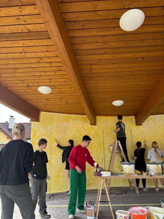 Foto vor einer gelben Wand mit Pinseln in der Hand. Im Vordergrung gelbe Farbkübel.