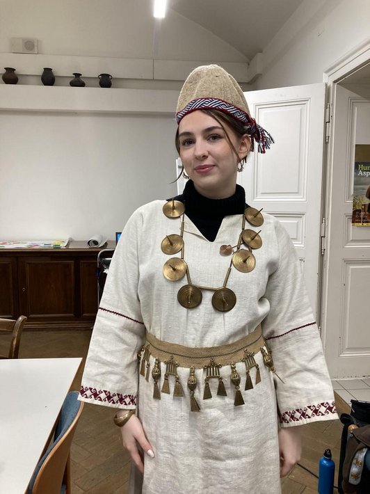 Foto einer Schülerin mit einem selbst genähten beigen Leinenkleid und Goldemen großen runden Haslkette und goldenem Gürtel, sowie einem genähten Hut.