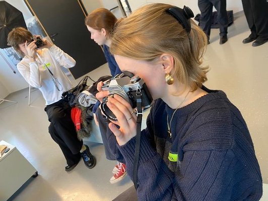 Nahaufnahme einer Schülerin mit einer analogen Kamera vor dem Auge, im Hintergrund eine weitere Schülerin mit Kamera, die eine sitzende Schülerin fotografiert.