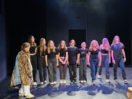 9 Schülerinnen auf einer Bühne nebeneinander stehens. Ein Mädchen geht mit einem Tiegermantel vorne vorbei.