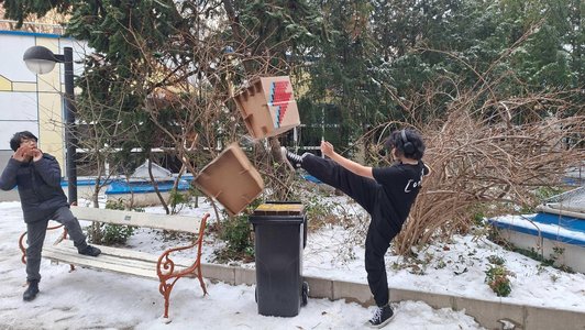 Foto von zwei Schülern im Wintre in einem Park. Einer fotografiert, der ander kickt mit dem Fuß einen Hocker aus Karton mit einem Blitz drauf.