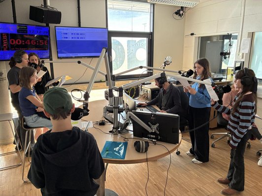 Foto von sieben Jugendlichen in einem Radiostudio vor Mikrophonen und Textnotitzen.