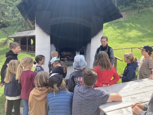 mehrere Schüler/innen stehen vostehen vor Holzofen und Frau mit Schürze