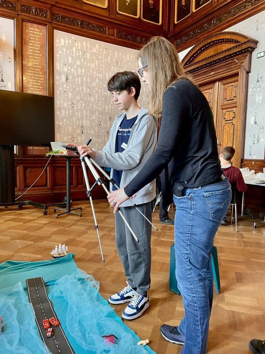 Schüler Fotografiert auf Boden arrangierte Szene, Frau daneben hält mit ihm Stativ