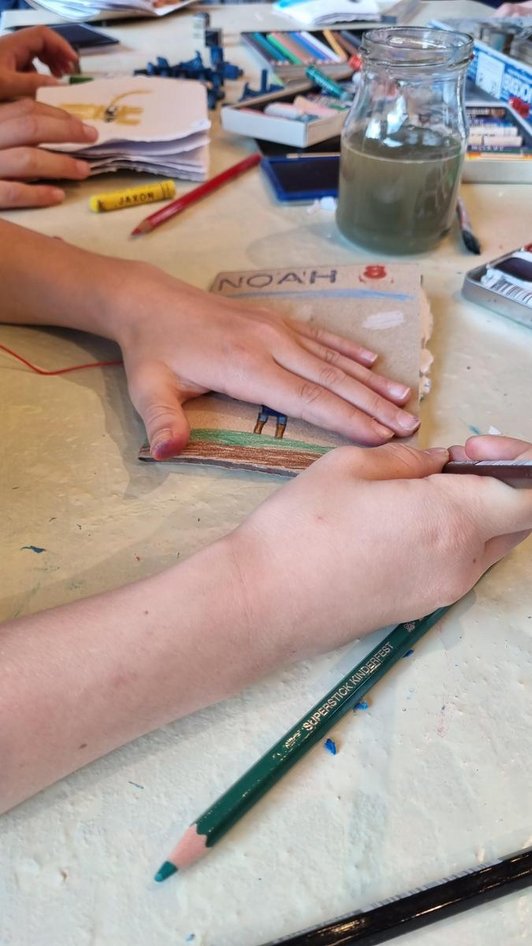 Tisch voller Stifte und Farben, Hände beim Gestalten selbst gebundener Heftchen