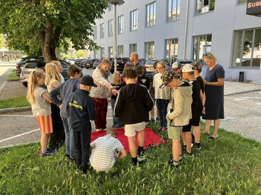 Schulklasse und drei Erwachsene stehen um rotes Tuch im Gras