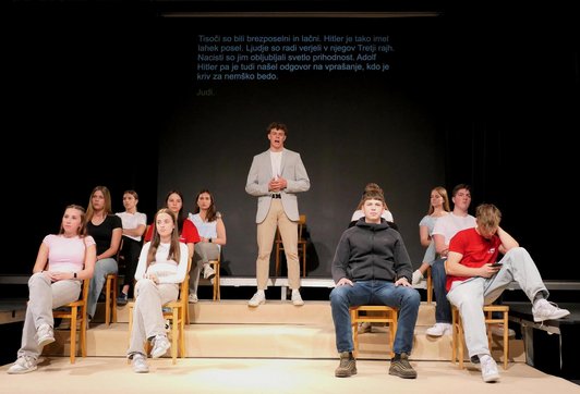Schüler sitzen auf einer Stiege auf einer Bühne und schauen ins Publikum. Ein junger mann steht im Mittelpunkt und doziert.