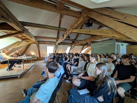 Mehere Schulklasse sitzen in einem Veranstaltungsraum und hören einem Interview zu, das auf einem Podium davor stattfindet. 