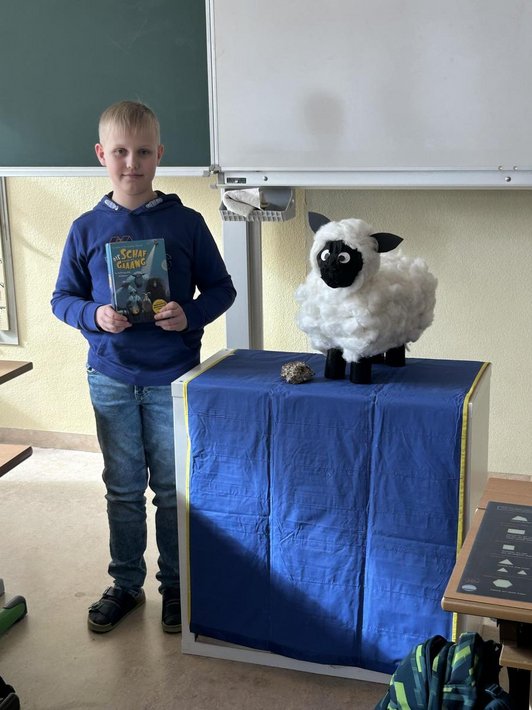 Foto von einem Schüler vor einer Tafel mit einem Buch Titel "Die Schafgaaang" in der Hand. Neben ihm auf einem Pult ein gebasteltes Schaf.