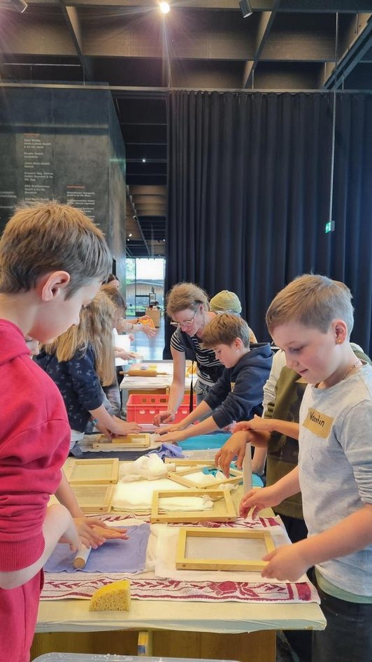 An einem langen Tisch schöpfen Kinder Papier mit einem Schöpfrahmen