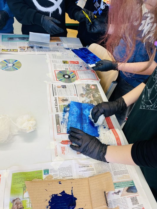 Foto von drei Schülerinnen und Schülern, die schwarze Gummihandschuhe tragen und Druckstöcke aus Tetrapaks blau einfärben