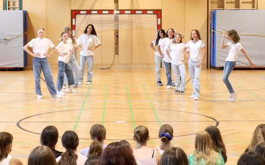 Neun Mädchen in Jeans und weißen T-Shirts tanzen in einer Linien in einem Turnsaal.