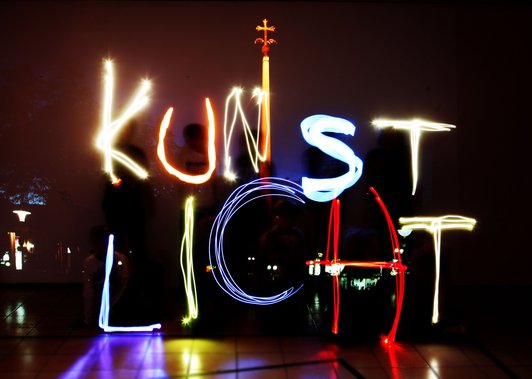 Foto von einer Bühnensituation, in der das Wort ''Kunstlicht'' mit einer Lichtquelle gezeichnet wurde