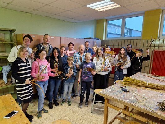 Foto von Schüler/innen und Lehern und Großeltern in einem Werksaal.Sie halten selbstgemachte Objekte aus Weide in ihren Händen