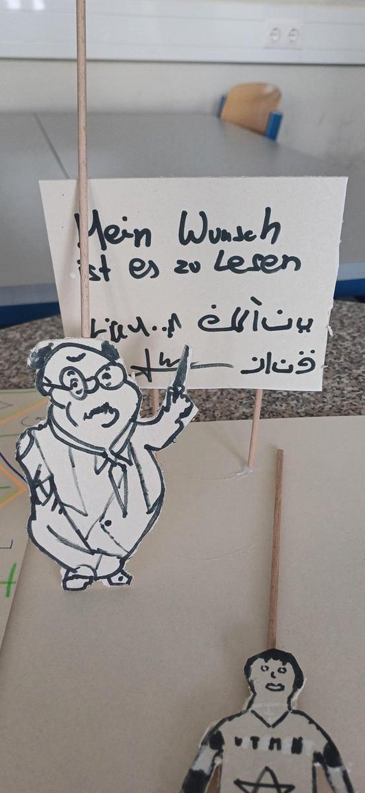 Auf stecken geklebte kleine Figuren und Tafel mit Text auf Deutsch und Arabisch
