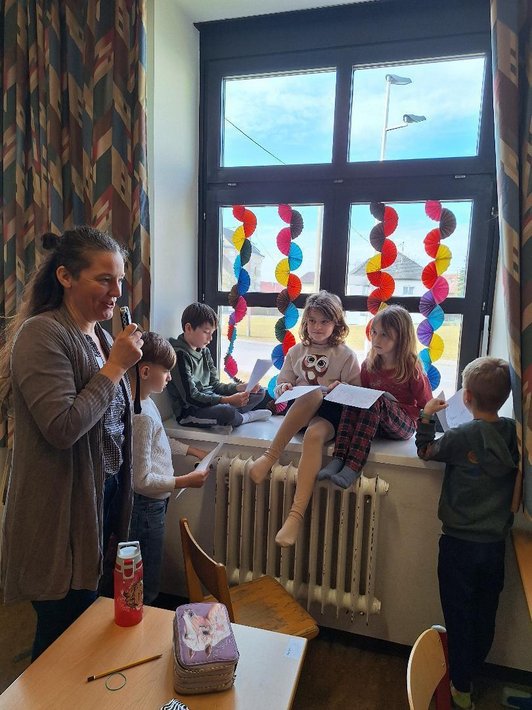 fünf Kinder mit Papierblättern in Händen sitzen auf und stehen an Fensterbrett neben Frau