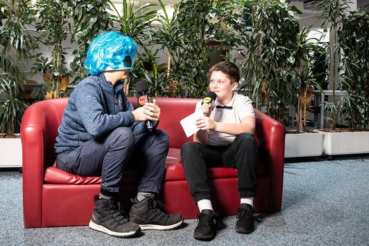 zwei Schüler mit Mikros auf roter Couch, einer mit blauer Maske