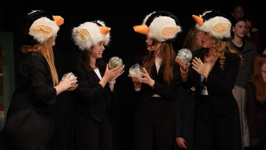 Szenenausschnittvon vier Schülerinnen auf einer Bühne alle schwarz gekleidet mit Hüten in Schwaneneform und einer durchsichtigen gefüllten Kugel in der Hand.