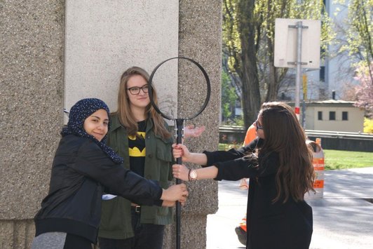 Foto von drei Schülerinnen auf dem Friedrich-Engels-Platz, die eine überdimensionale Lupe halten