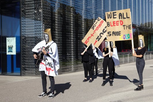 Foto von sechs Lehrlingen mit Masken und zwei Transparenten bei der Performance neben dem Lentos 