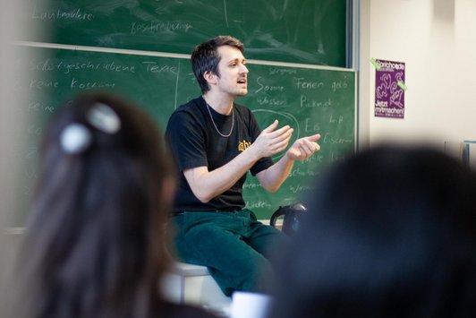 Mann sitzt gestikulierend mit geöffnetem Mund auf Tisch vor Schultafel, zwei Köpfe im Vordergrund 