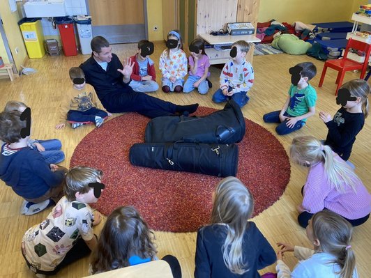 Mann sitzt mit Kindern um runden Teppich auf dem zwei Intrumententaschen liegen