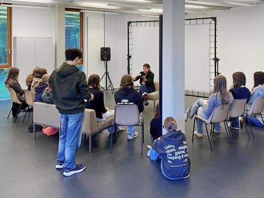 Schüler/innen sitzen in Halbkreis um gestikulierende Frau