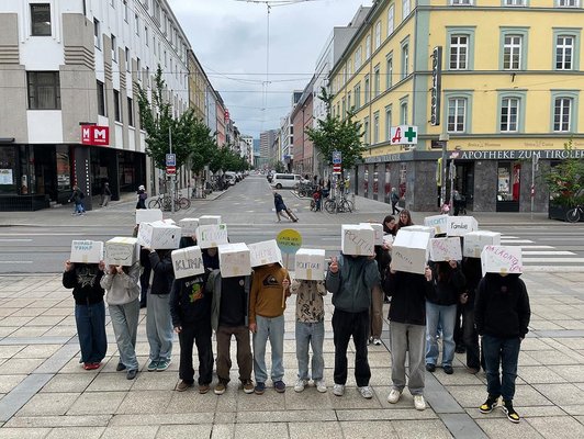 Schüler/innen mit beschrifteten Boxen überm Kopf stehen zur Kamera gewandt vor Straße