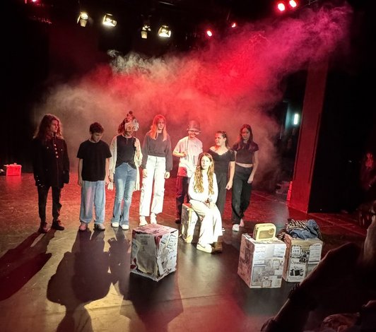 Foto von Schülerinnen auf einer Bühne mit Boxen beklebt mit Zeitungspaperi und Nebel.