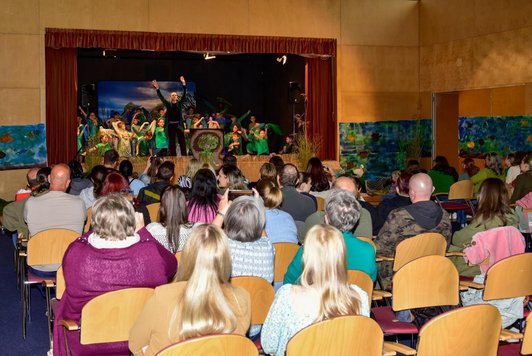 Schüler/innen in grünen T-Shirts und Frau auf Bühne, davor Erwachsene in Sesselreihen