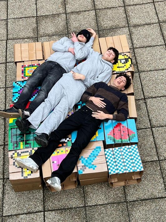 Foto der fertigen bunten Hocker aus Karton zusammengestellt. 3 Schüler liegen auf ihnen von oben fotografiert..