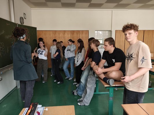 Foto von Schüler:innen in einer Klasse stehend und sitzend vor der Tafel.